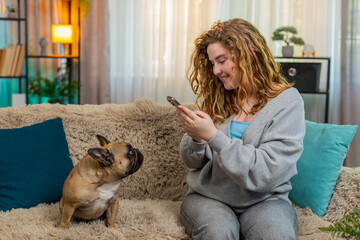 Young Caucasian redhead woman smiles while using smartphone to take photo of pug dog sitting on...