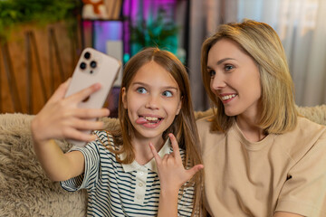 Young child girl sitting with mother on home sofa, posing for selfie and making silly faces laughing out loud together. School kid and mom enjoy joyful bonding moment while using smartphone for photos