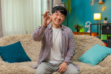Asian man sitting on home sofa smiles at camera, giving OK hand gesture, positive assurance...