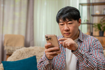 Asian man on home sofa fills online form on smartphone, carefully tapping screen focused serious...