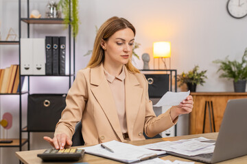 Middle-aged businesswoman at home office sorts receipts uses calculator balances budget reviews...