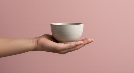 Hand holding a small ceramic bowl with a smooth surface against a soft pink background delicate product presentation.