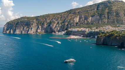 Fototapeta premium Aerial photo of Meta di Sorrento and Piano di Sorrento seen from the sea, with the Sorrento coast shining under the summer sun. A stunning seaside view of cliffs, beaches, and blue waters. Italy.