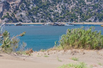 Obraz premium A landscape image showing calm waters near a mountain. There is vegetation in the foreground, with possible fish farms visible on the water on Turtle Beach - Dalyan Delta - Dalyan - Mugla - Turkey