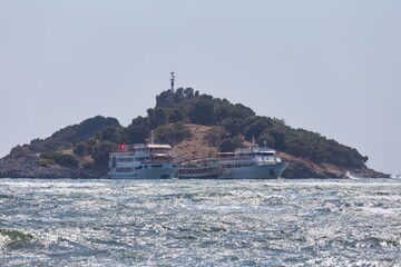 Fototapeta premium Two boats rest along the coast of a small island, covered in trees, under a cloudless blue sky. A small structure sits atop the peak of the island on Turtle Beach - Dalyan Delta - Dalyan - turkey