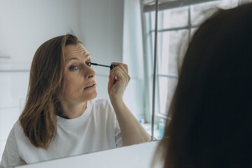 Middle-aged woman applying eye makeup with a brush in front of a bathroom mirror. Beauty routine...