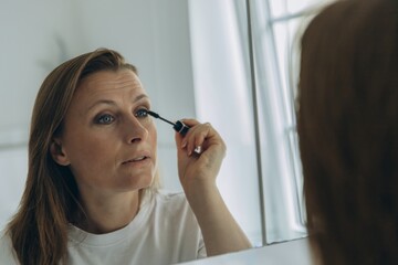 Middle-aged woman applying eye makeup with a brush in front of a bathroom mirror. Beauty routine...