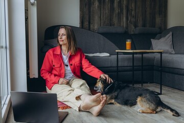Freelancer woman working from home with laptop, sitting on floor in cozy living room, barefoot and...