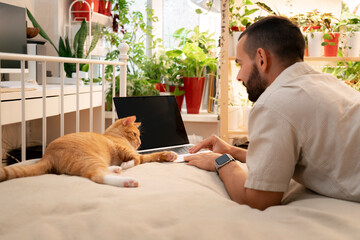 A man is typing on his laptop while lying on his stomach on a bed. An orange tabby cat is lying...