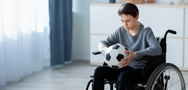Full length of teen football player in wheelchair holding soccer ball and feeling stressed over his injury at home. Adolescent sportsman suffering from depression over his disability