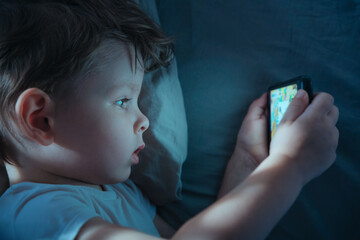Close-up of small boy lying in bed at night, eyes wide while watching cartoons on smartphone or...