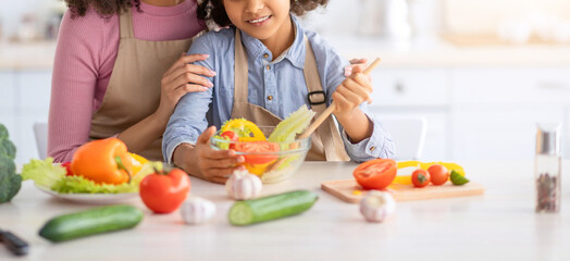 Healthy Food And Diet Concept. Portrait of happy black family cooking tasty meal together in the kitchen, charming girl mixing salad in bowl, black woman embracing her and teaching how to cook