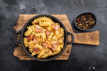 Fusilli carbonara pasta topped with crispy bacon bits and parmesan  cheese  in a skillet. Black background. Top view