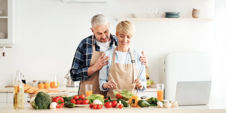 Enjoy of cooking healthy food together, modern blog and love. Happy middle aged european man hugs smiling wife making salad in modern kitchen interior with bright fresh vegetables on table with laptop