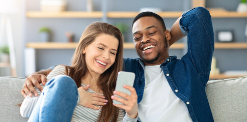 Cheerful Interracial Lovers Watching Funny Videos On Smartphone At Home, Happy Multicultural Couple...