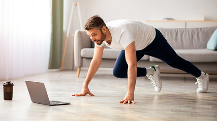 Positive athletic bearded middle-aged man exercising at home, using laptop, watching sport videos...