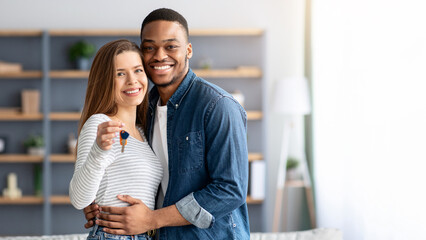 Happy young multiracial couple holding home keys and hugging, romantic millennial family, black man and white woman poising in living room after moving to new appartment, smiling at camera, free space