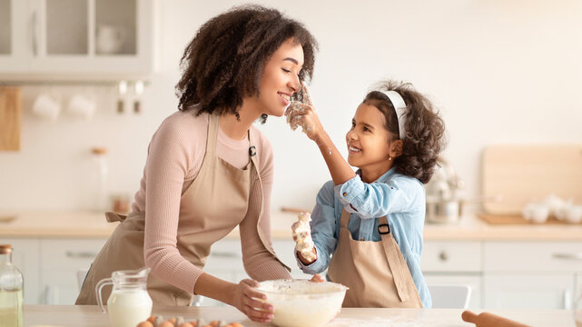 Cooking With Family Is Fun. Portrait of happy African American mom and her daughter baking in kitchen and fooling around, girl playfully touching woman's nose, kneading dough and having fun
