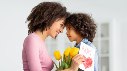 Happy Loving Family. Portrait of African American woman and little girl touching foreheads, looking at each other, daughter greeting mommy with mother's day, lady holding card and bunch of flowers