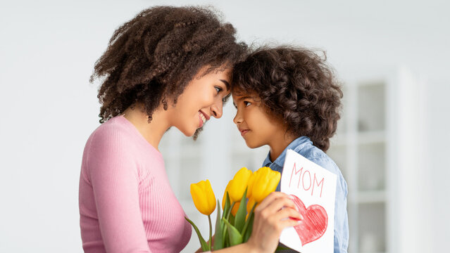 Happy Loving Family. Portrait of African American woman and little girl touching foreheads, looking at each other, daughter greeting mommy with mother's day, lady holding card and bunch of flowers - Powered by Adobe