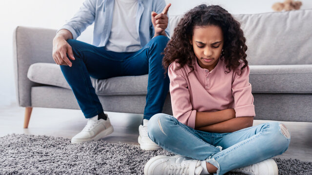 Naughty Kid. Portrait Of Angry Annoyed African American Father Scolding His Upset Little Daughter For Her Behaviour While She Sitting On The Floor At Home. Family Conflicts And Punishment Concept - Powered by Adobe