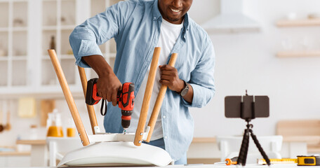 Modern blogger repairman shooting video. African american man making chair with electric drill,...