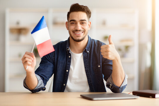 Smiling arab guy with flag of France showing thumb up, sitting at desk with laptop, middle-eastern young man student learning French for job, education or emigration to France, copy space