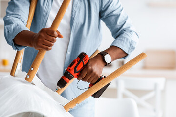 Furniture assembly process. African american handyman screwing legs to chair with electric...