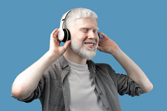 Happy albino man in headphones listening to music, enjoying his favorite song on turquoise studio background. Positive guy with unusual appearance in headset relaxing to beautiful melody - Powered by Adobe