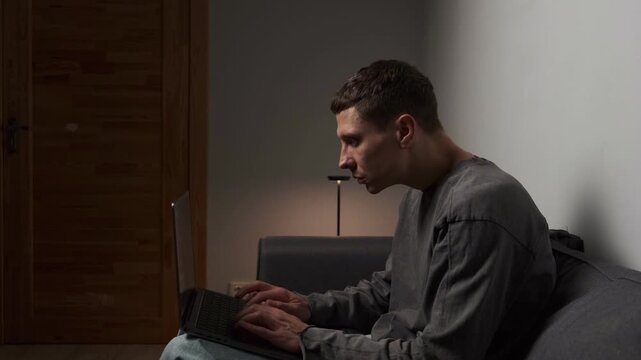 Incorrect posture while working on laptop. Young man sitting hunched on sofa using laptop with poor ergonomics, harmful effect on spine and posture in authentic modern living room