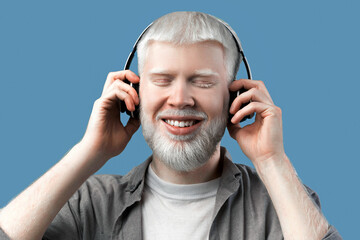 Happy albino man listening music in wireless headphones and touching them on turquoise studio background. Peaceful guy closing eyes and smiling, enjoying favourite melodies