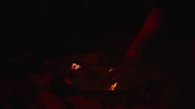 Person stirs embers in night campfire with wooden stick