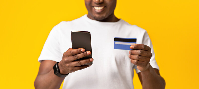 Shopping with pleasure, online order and application. Cheerful millennial african american man in white t-shirt uses smartphone and credit card, isolated on orange background, studio shot, cropped