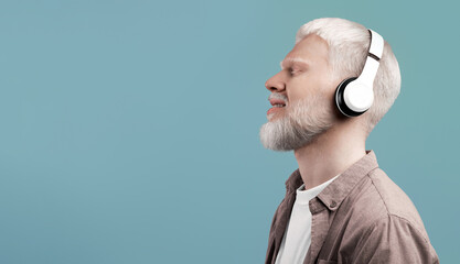 Enjoy the sound. Peaceful albino man listening to music with closed eyes in wireless headphones. Guy enjoying favourite melodies with pleasure and smiling, grey background, copy space