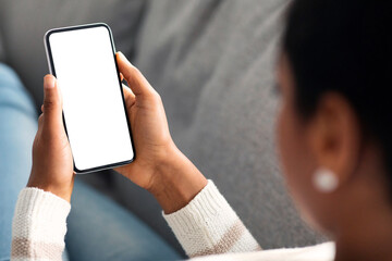 Unrecognizable Black Woman Holding Smartphone With White Screen At Home, Over Shoulder View Of African American Lady Using Blank Mobile Phone While Relaxing On Couch, Mockup Image With Copy Space