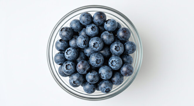 Overhead shot of fresh blueberries in a clear glass bowl on a white surface in bright light