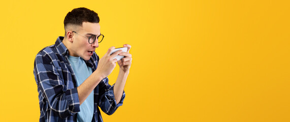Online gaming. Addicted young guy playing video games on cellphone, overusing his gadget on orange studio background. Gamer using smartphone for entertainment and fun