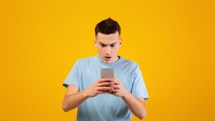 Handsome young guy staring at mobile phone in shock over orange studio background. Cool millennial...