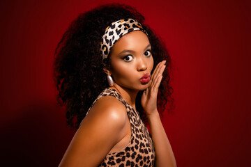 Tempting young woman in leopard print outfit with curly hair and headband posing against red...