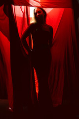 Silhouette of a woman standing gracefully behind red fabric in a dimly lit studio environment...