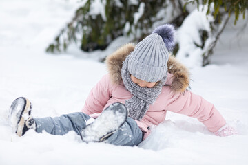 Happy excited little girl in warm clothes sitting on a snow, happily playing in winter park and...