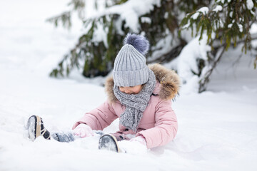 Happy excited little girl in warm clothes sitting on a snow, happily playing in winter park and joyfully laughing. Fun outdoor activities for children in wintertime. Winter portrait with copy space