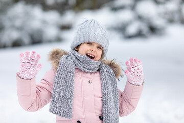 Portrait of happy excited little girl in warm pink and grey clothes playing in snowy park, joyfully...