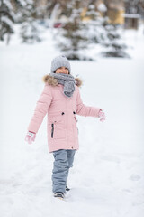 Happy little girl in warm pink and grey outfit walking on the street in winter park, joyfully...