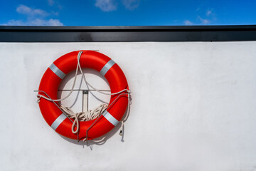 Bright red lifebuoy with attached rope mounted on a white wall against blue sky background,...