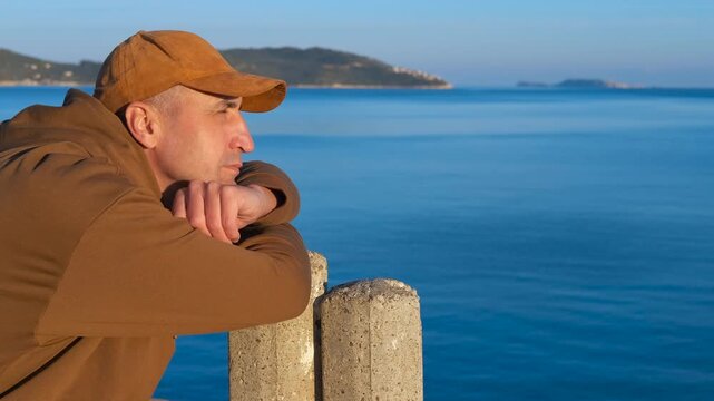 A thoughtful man enjoys the seascape. Pensive adult man wearing brown baseball cap and casual jacket leaning against concrete bollard, gazing pensively at tranquil blue seascape with distant horizon