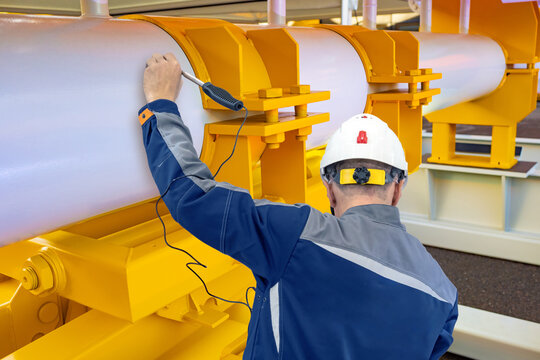 Engineer in hard hat performs ultrasonic testing on industrial pipeline using inspection equipment. Activity: Nondestructive Testing, Engineering, Industrial Inspection, Maintenance.