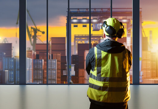 Worker at container port. Man in customs warehouse terminal. Supervisor stands with his back to camera inside customs port with panoramic windows. Man in vest looks at gantry crane with containers