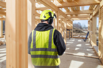 Construction worker in safety vest and helmet inspects wooden frame of building under construction,...