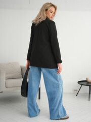 Stylish Woman in Oversized Blazer and Wide-Leg Jeans Poses Confidently in Modern Interior Setting...